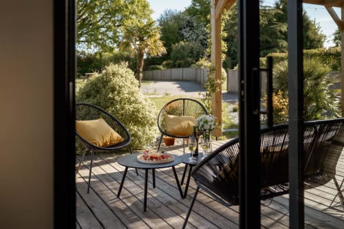 Shaded terrace at Manoir de la Nogrie cottage with wicker chairs, round table, and flowering garden view