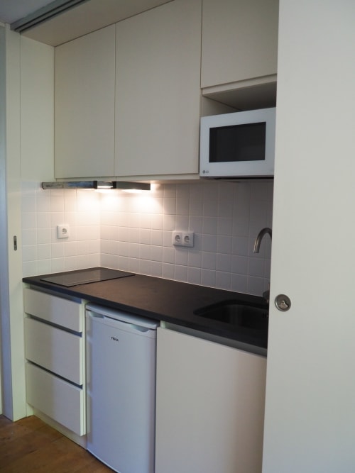 Cozinha compacta moderna com armários brancos, bancada preta, micro-ondas e geladeira no apartamento Quinta da Coutada