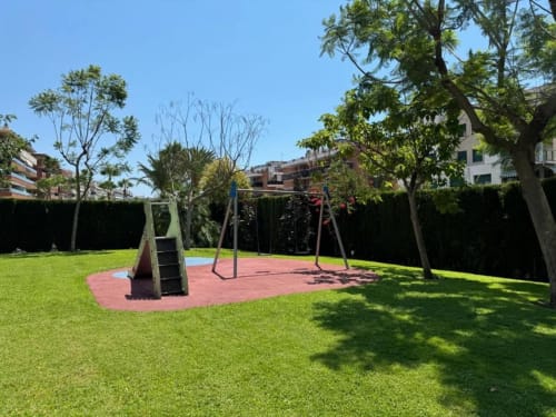 Sunny garden area at Rentalaya apartment in Salou with playground equipment, green lawn, and surrounding hedges