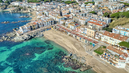 Aerial view of Palafrugell coastal town with turquoise Mediterranean waters, sandy beach, and colorful waterfront buildings.