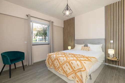 Quarto moderno com cama de casal, manta laranja, poltrona azul-petróleo, luminária geométrica e janela com vista para a piscina