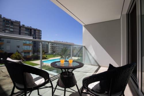 Varanda ensolarada de apartamento com vista para piscina em Rent-A-Key Portugal, com cadeiras de vime preto e bebidas