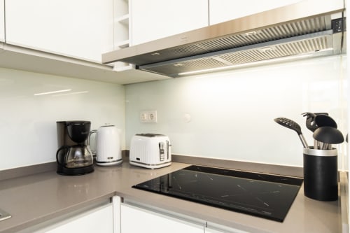 Cozinha de apartamento moderno com armários brancos, cooktop indução, eletrodomésticos de aço inoxidável e suporte preto para utensílios