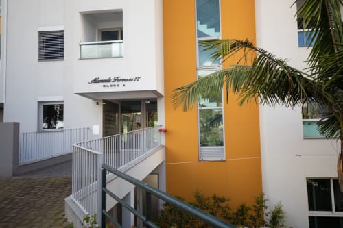 Entrada de edifício moderno com fachada branca e laranja, palmeiras, guarda-corpos metálicos e acessibilidade no nível do solo