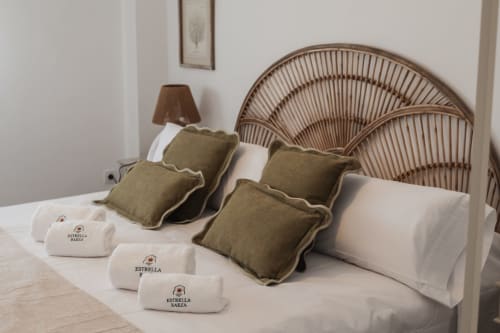 Bedroom with woven rattan headboard, olive green pillows, white linens, and Estrella Baeza amenities at luxury apartment in