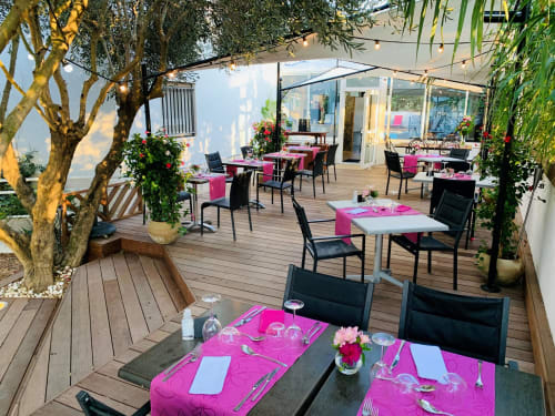Terrasse Entre 2 eaux in Marseillan-Plage mit rosa Tischdekorationen, schwarzen Stühlen, Bäumen und Seeblick