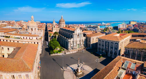 Vista aerea della Piazza del Duomo di Catania con chiese barocche, tetti in cotto e il Mar Mediterraneo sullo sfondo