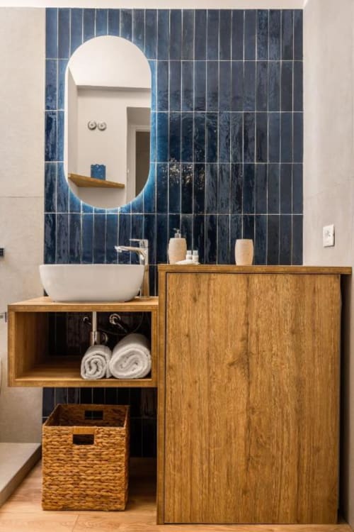 Modern bathroom with illuminated oval mirror, white vessel sink, wooden vanity storage, rolled towels, and dark blue tile