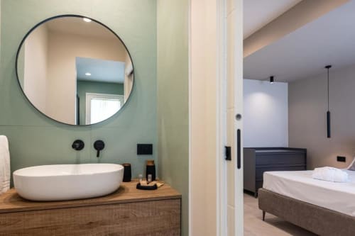 Bagno moderno con specchio rotondo e lavabo bianco, camera da letto adiacente con letto matrimoniale presso la villa Dimora Silva Nature Suites