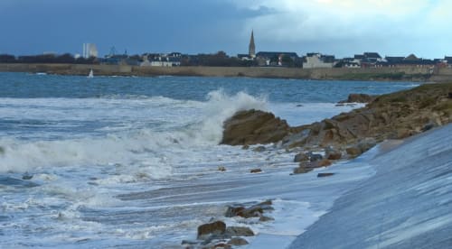 Côte rocheuse de Normandie avec vagues et silhouette de Saint-Pair-sur-Mer à l'horizon