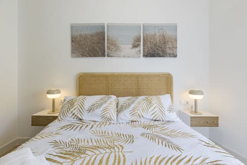 Dormitorio moderno con cabecero de ratán, ropa de cama con patrón de pan de oro, mesitas con lámparas y arte de dunas en Numidas