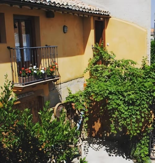 Acogedora casa rural española amarilla con balcón de hierro forjado, macetas floridas y frondoso jardín en el patio de Casas