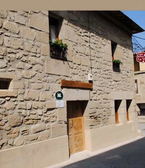 Fachada de piedra de la casa rural La Viña en Cuzcurrita de Río Tirón con puerta de madera y macetas de flores en ventanas