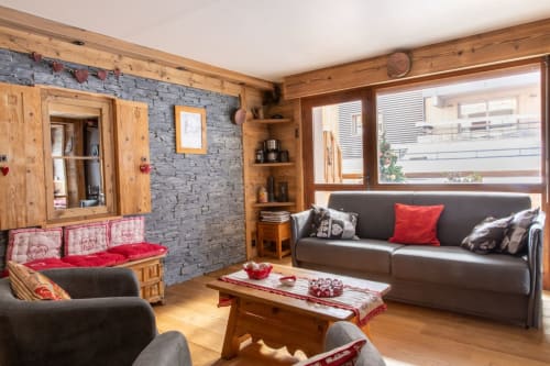 Salon cosy Zoppas Courchevel avec poutres en bois, mur en pierre, canapés gris et accents rouges