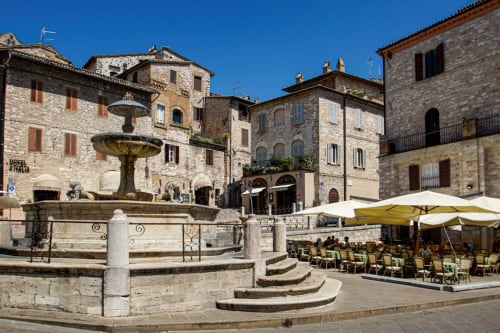 Storica piazza in pietra ad Assisi con fontana, edifici medievali e tavoli all'aperto dell'hotel Proprietà Clara