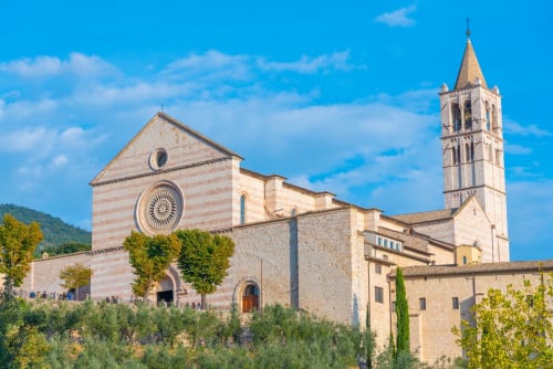 Basilica di Santa Chiara ad Assisi con campanile, rosone e cipressi verdi sotto il cielo azzurro