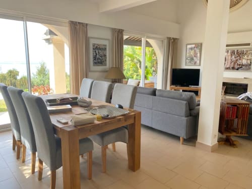 Luminoso soggiorno con tavolo da pranzo, divano grigio, TV e finestre con vista mare presso la Conciergerie De Porticcio in Corsica