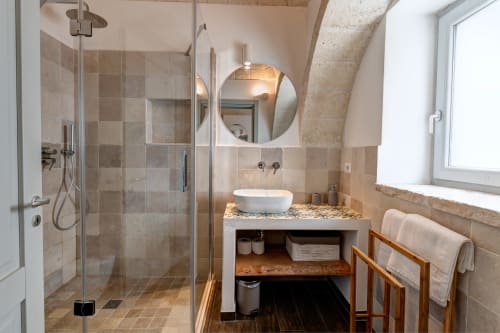 Bagno lussuoso nella villa Trulli Gianpaolo a Bari, con finiture in pietra, doccia walk-in, specchio rotondo e lavabo a bacinella