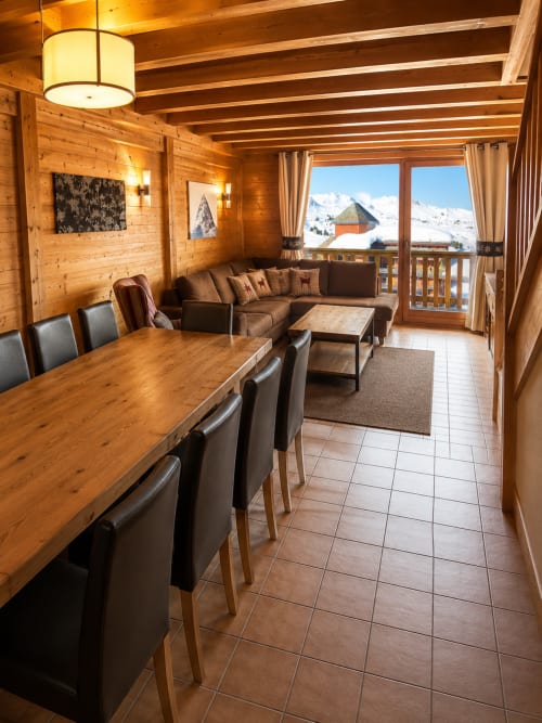 Spacieux séjour alpin avec poutres en bois, table à manger, canapé marron et vue panoramique sur les montagnes enneigées depuis le balcon du