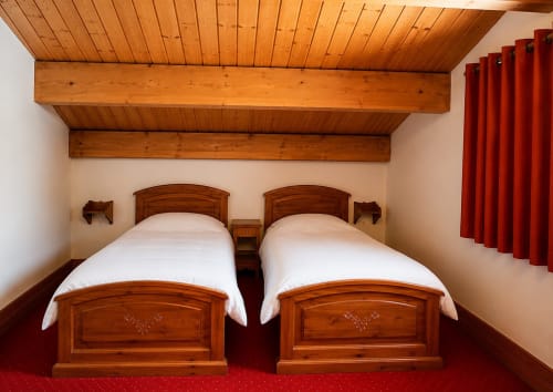 Deux lits jumeaux aux cadres en bois dans une chambre lumineuse du Chalet des Cimes, Savoie, avec poutres apparentes et rideaux rouges