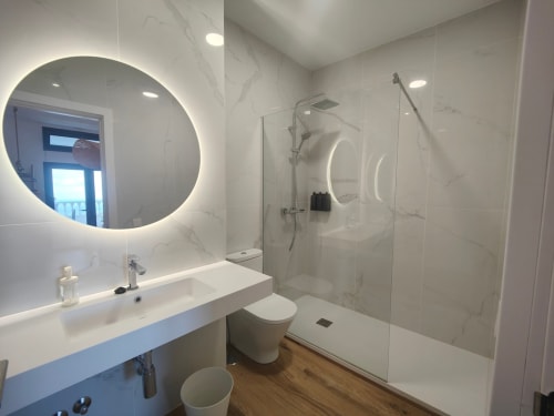 Modern bathroom at Nido Anaga with illuminated circular mirror, marble tiles, glass shower, and wooden flooring