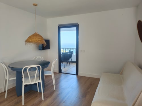 Comedor moderno en Nido Anaga con mesa azul, lámpara colgante tejida y vistas al mar a través de puertas de cristal hacia la terraza.