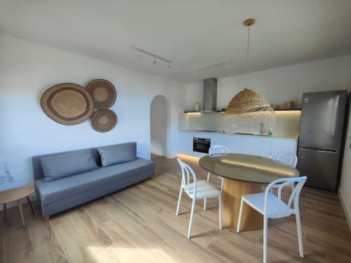 Apartamento moderno diáfano en Nido Anaga con mesa de comedor, sofá gris, cocina equipada y platos tejidos decorativos en la pared.