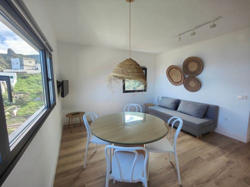 Apartamento moderno en Nido Anaga con ventanal de vista panorámica al mar, mesa de comedor, sillas azules, sofá gris, lámpara colgante tejida y
