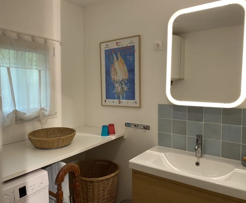 Salle de bain moderne de la villa Le Mas Bello Visto à Lorgues avec miroir illuminé, lavabo blanc, carrelage gris et accessoires en rotin