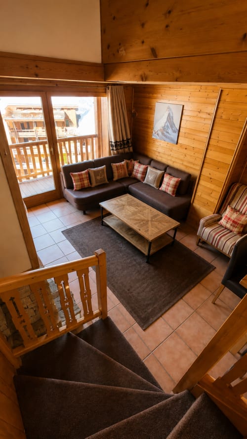 Salon d'appartement alpin avec vue sur les montagnes, canapé en cuir noir, murs en bois, accès au balcon et mobilier moderne