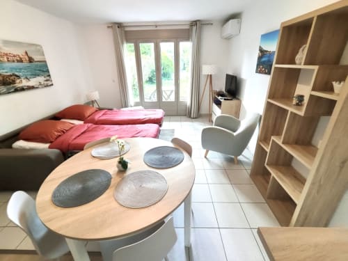 Appartement moderne lumineux à Barcaresa Port Leucate avec lit rose, table à manger, étagères en bois, TV et balcon avec vue sur le jardin