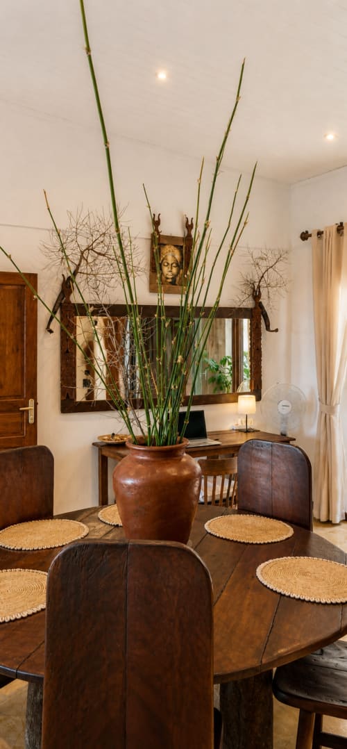 Sala da pranzo con tavolo in legno, sedie in pelle e vaso di rame con steli verdi alti presso La Villa Boraha, Vohilava
