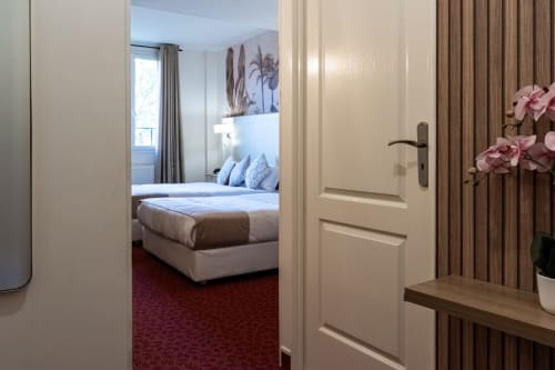 Ingresso della camera dell'Hotel Royal Paris Ivry con porta beige, tende in legno, fiori rosa e letto matrimoniale visibile dall'ingresso
