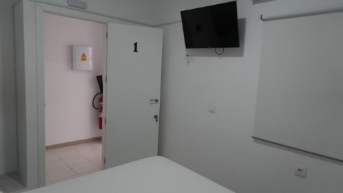 Habitación del Hostal Merino Jédula con paredes blancas, escritorio, TV mural, puerta a habitación contigua, extintor y pizarra