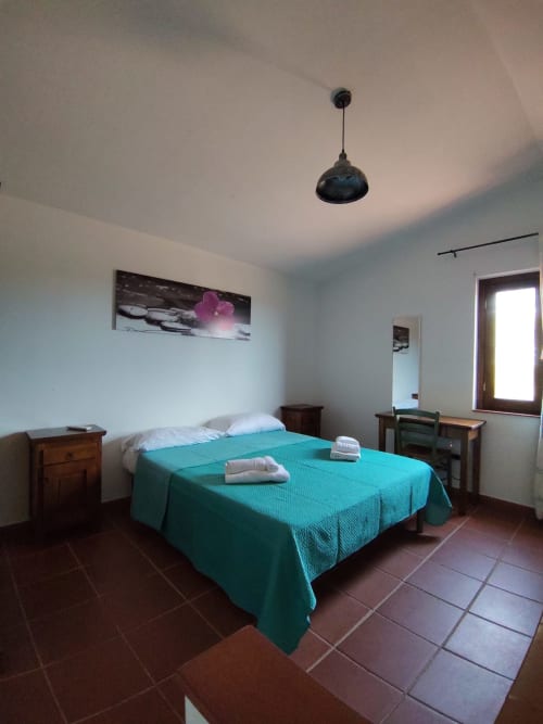 Bright bedroom at San Teodoro hotel with turquoise bedcover, wooden furniture, pendant light, and terracotta flooring