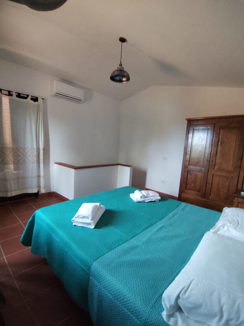 Spacious bedroom with turquoise bedcover, white towels, terracotta floor, wooden wardrobe, and modern pendant light at