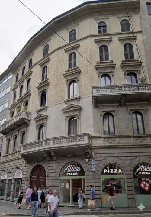 Historic Milano building facade with arched windows, ornate balconies, and ground-floor pizza shop on Via Torino street