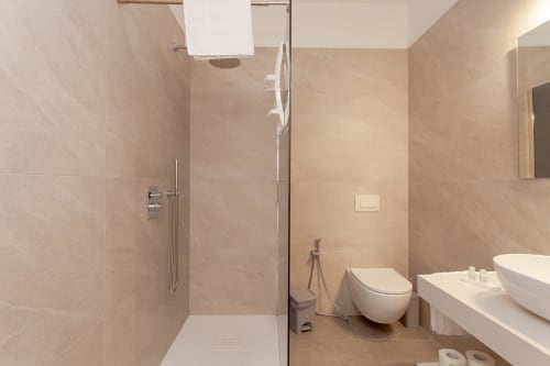 Modern bathroom at Le Dune Relax hotel with walk-in shower, wall-mounted toilet, dual sinks, and beige tile finishes in San