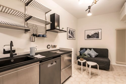 Angolo cottura moderno con elettrodomestici in acciaio inox e piani neri, collegato a zona living con divano scuro da Flavia's