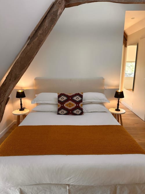 Chambre mansardée moderne avec poutre apparente, lit blanc, coussin motifs, lampes de chevet et lumière chaleureuse au Gîte