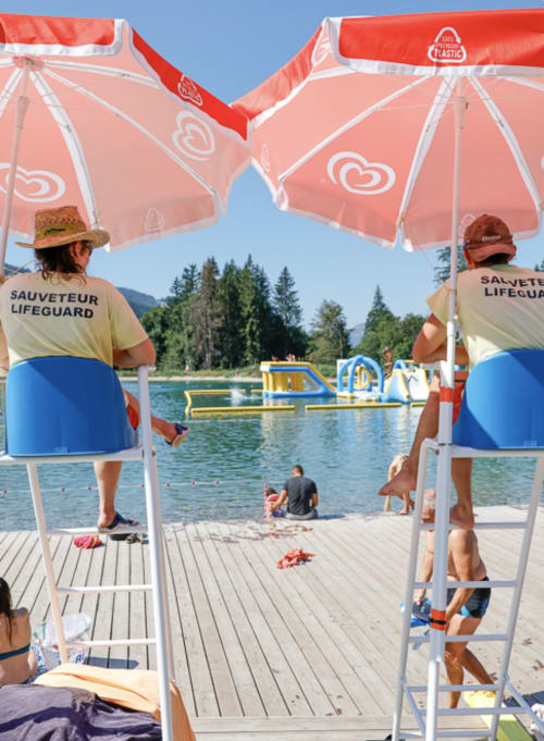 Lifeguards monitor swimmers at Chez Francine's beach, Flumet. Red umbrellas shade the sandy shore with lake and forest views.