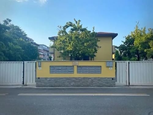 Ingresso di La Casetta dei Ronchi con facciata gialla e cancellata in metallo, circondato da alberi sotto cielo sereno