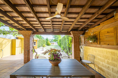 Terraza cubierta con vigas de madera, mesa de comedor y macetas de flores con vistas al jardín de la casa rural Ca L'Avi