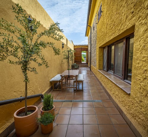 Terraza soleada de la casa rural Ca L'Avi con macetas, mesa de comedor de madera, muros de piedra amarilla y vistas al jardín