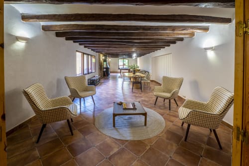 Amplio salón de la casa rural Ca L'Avi con vigas de madera vista, sillones de cuadros, alfombra redonda y suelo de terracota.