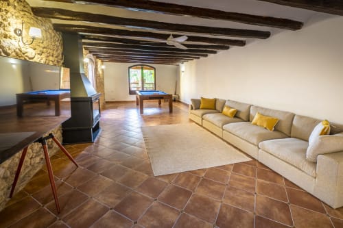 Amplia sala de juegos en la casa rural Ca L'Avi con mesas de billar, sofá beige con cojines amarillos, vigas de madera y vistas al jardín