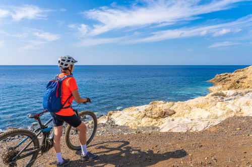 Ciclista con maglietta rossa e casco bianco su sentiero costiero sassoso con vista sul blu Mediterraneo presso La Casetta dei Ronchi