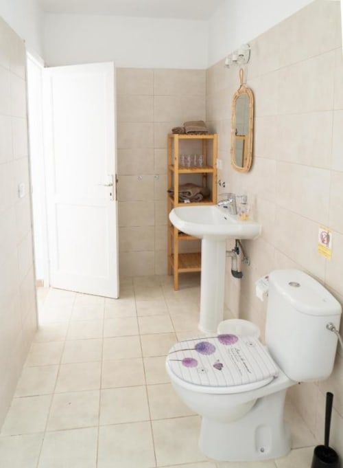 Clean bathroom in Villa Estrella Polar apartment with toilet, pedestal sink, wooden shelf, mirror, and white tile flooring.