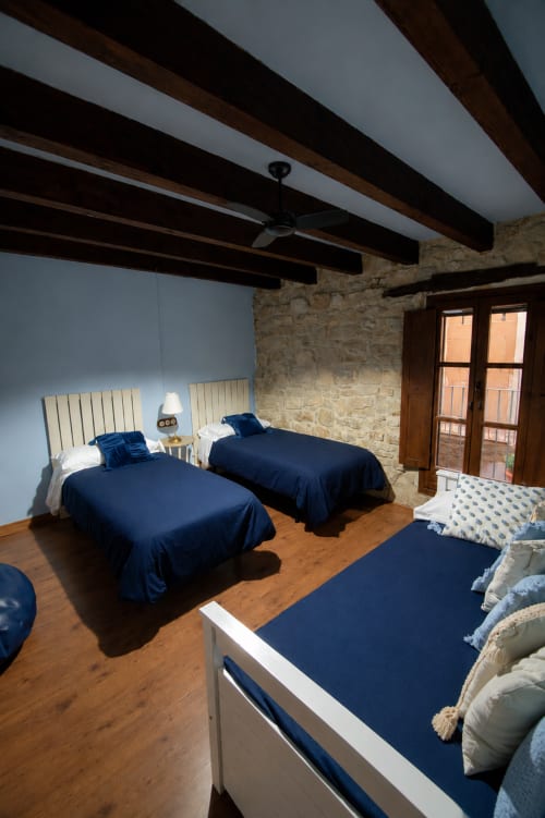 Habitación con múltiples camas en Cal París Rural, con ropa de cama azul marino, vigas de madera expuestas, muros de piedra y suelos de madera en La Donzell