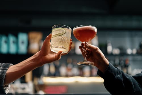 Due mani che brindano al bar La Casetta dei Ronchi, una con un drink trasparente, l'altra con un cocktail ambrato